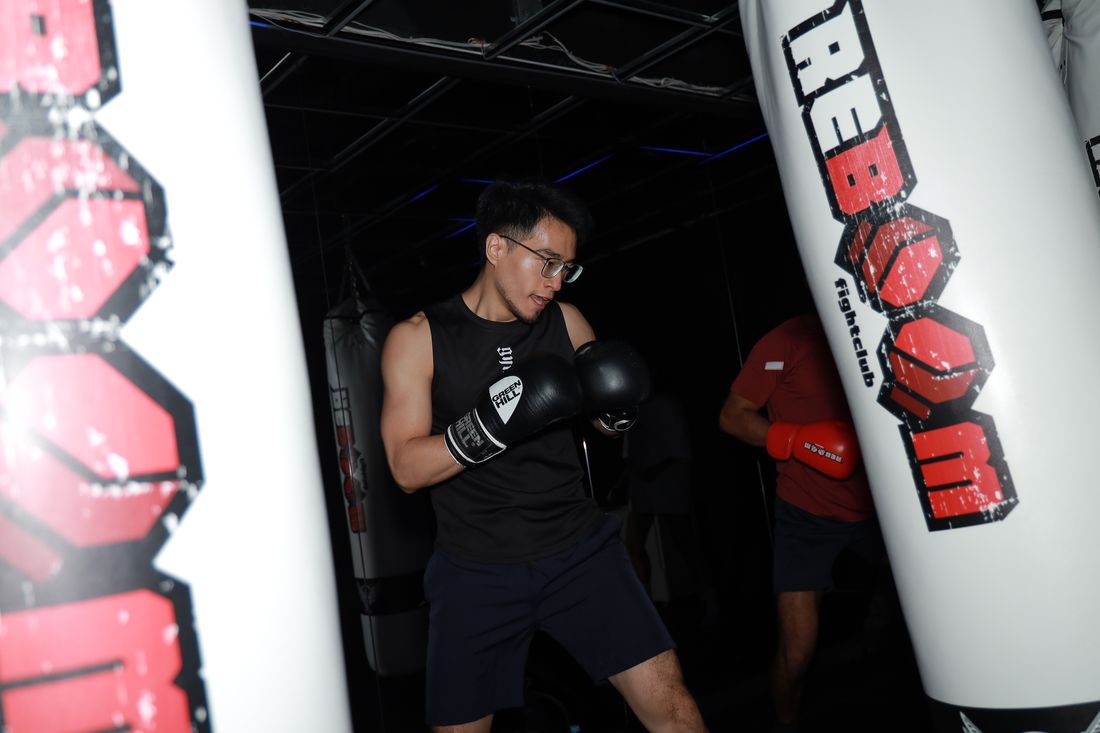 一位戴眼鏡的男生穿黑色背心，戴著拳擊手套在兩顆沙包之間出拳