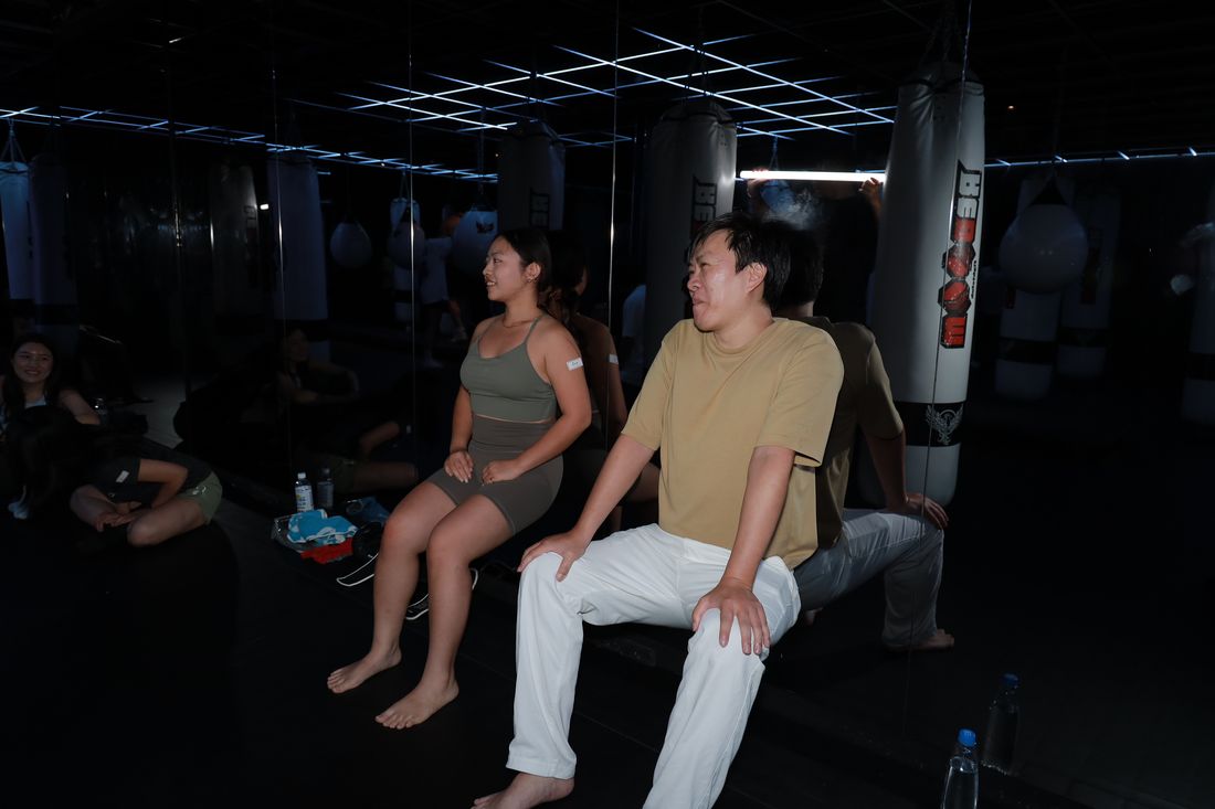 兩位參加者背靠著牆做靠牆半蹲，表情吃力，旁邊有人坐在地上看著