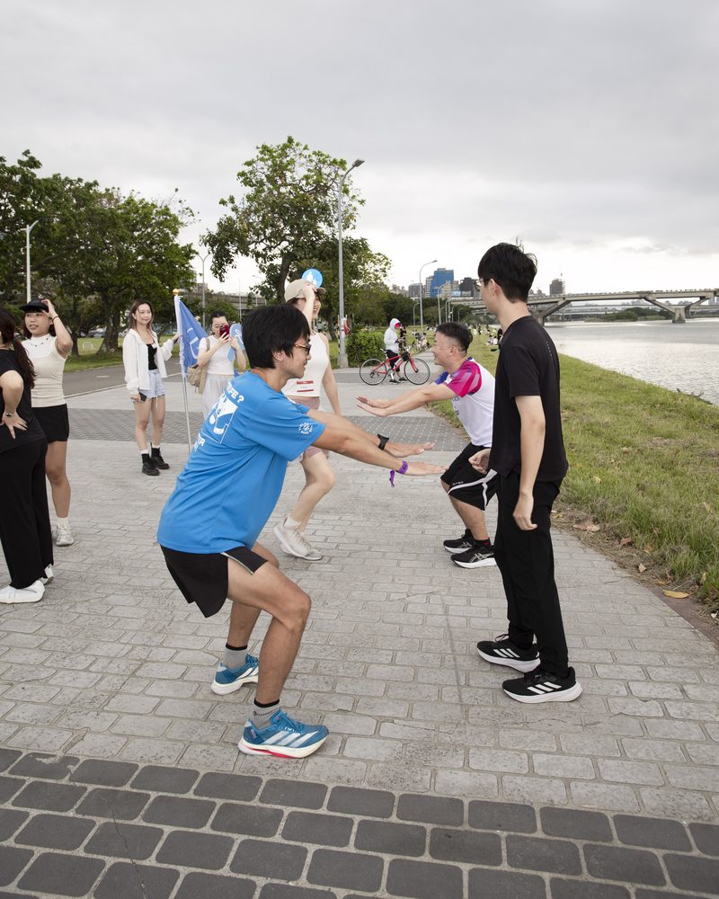 河濱步道上,兩位男生面對面拉著手做雙人深蹲暖身,後方是淡水河與其他正在暖身的參加者