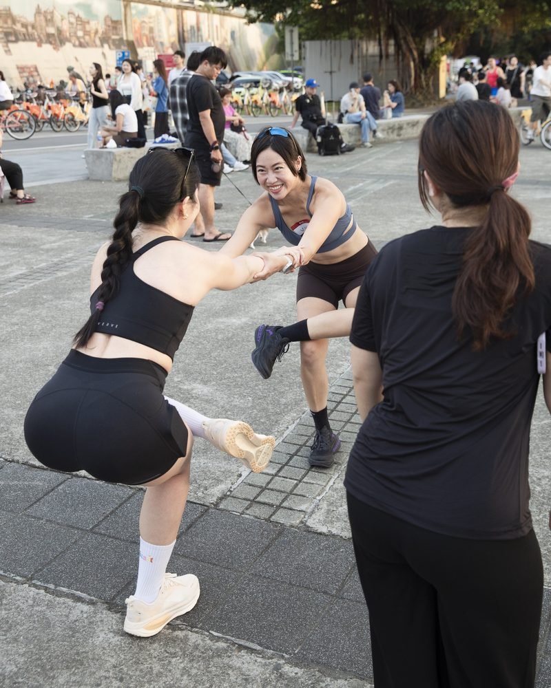 碼頭廣場上,兩位女生牽著手做單腳平衡伸展,其中一位笑得很開心,旁邊的人在看
