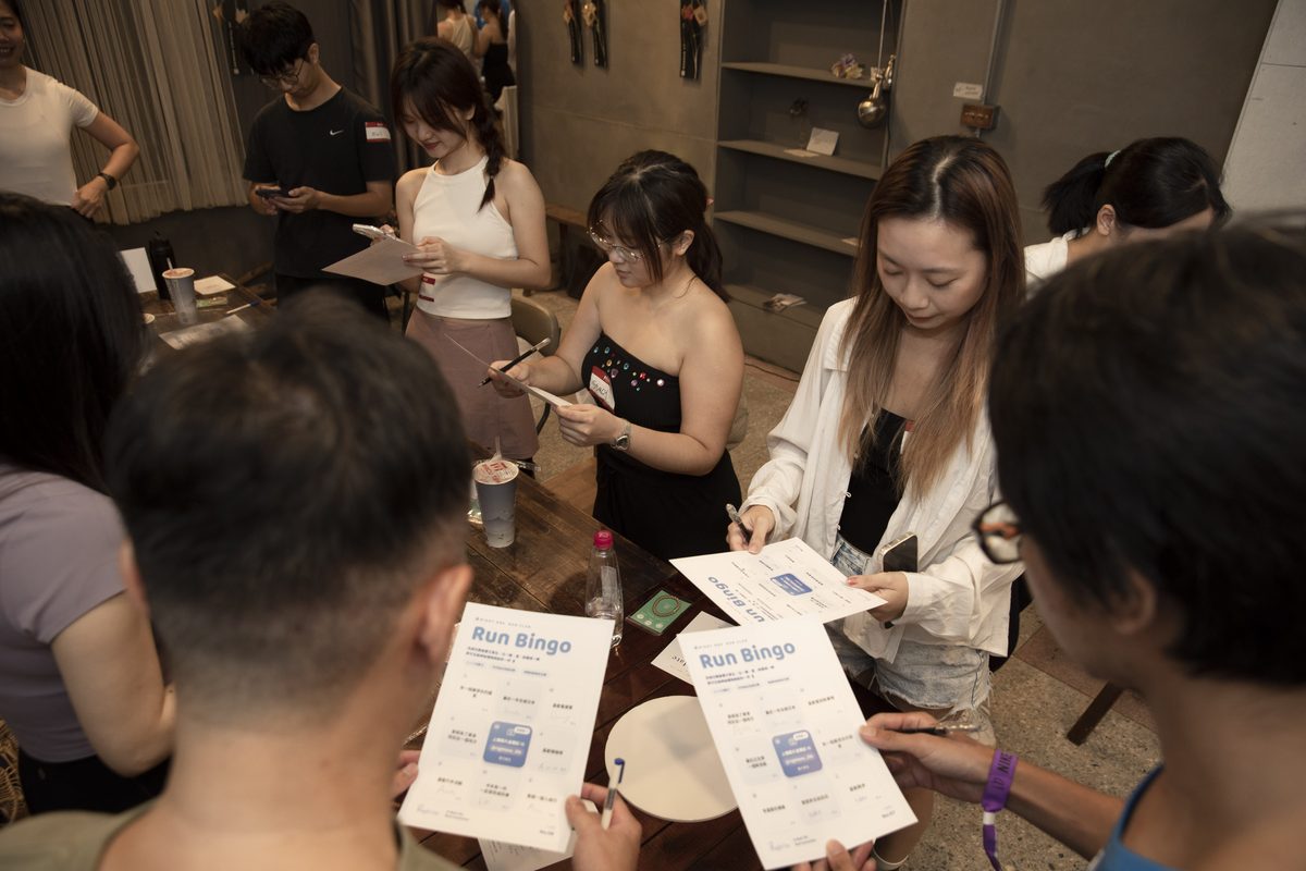 室內場地裡,一群參加者圍著木桌,手上拿著印有 Run Bingo 的賓果卡和筆,低頭互相對照格子