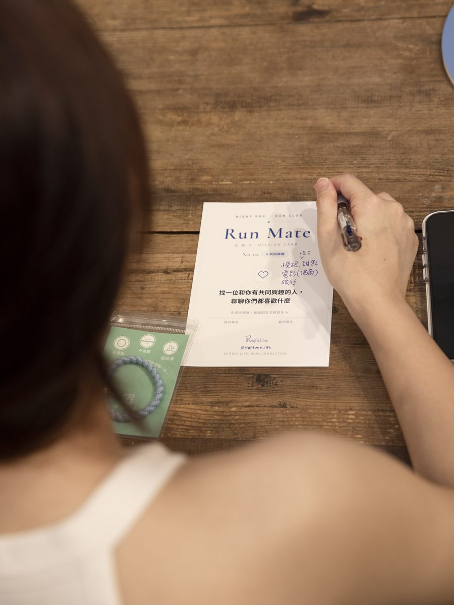木桌上放著一張 Run Mate 任務卡,寫著「找一位和你有共同興趣的人,聊聊你們都喜歡什麼」,一隻手正拿筆在卡上寫字