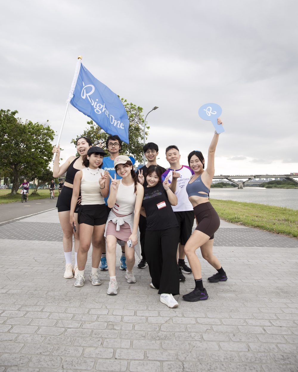 延平河濱的步道上，一群穿運動服的參加者舉著 Right One 藍色旗子擠在一起合照，背後是淡水河與陰天的傍晚天空