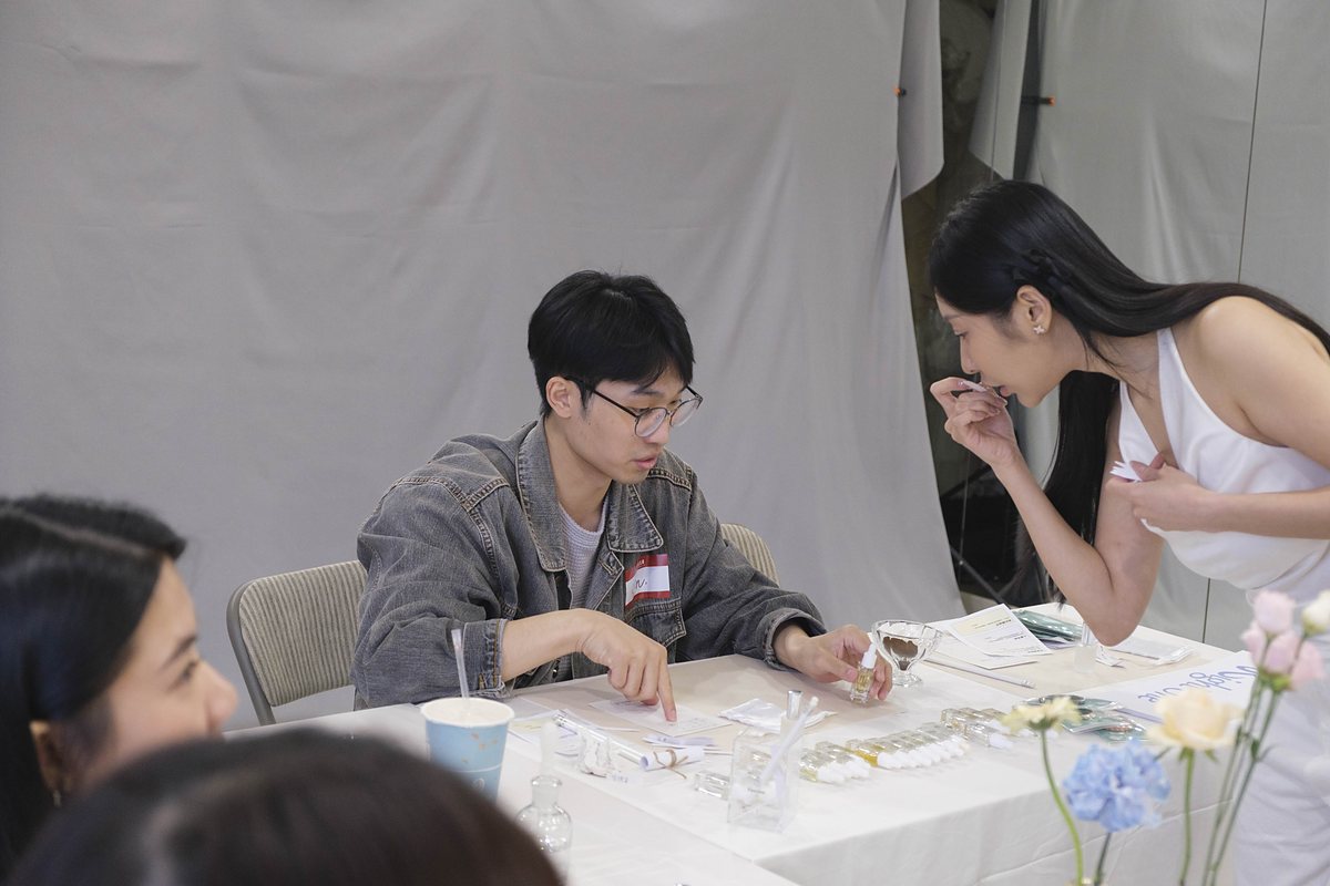 一位男生坐在桌邊調配香水,旁邊的女生彎腰低頭聞他手上的試香紙